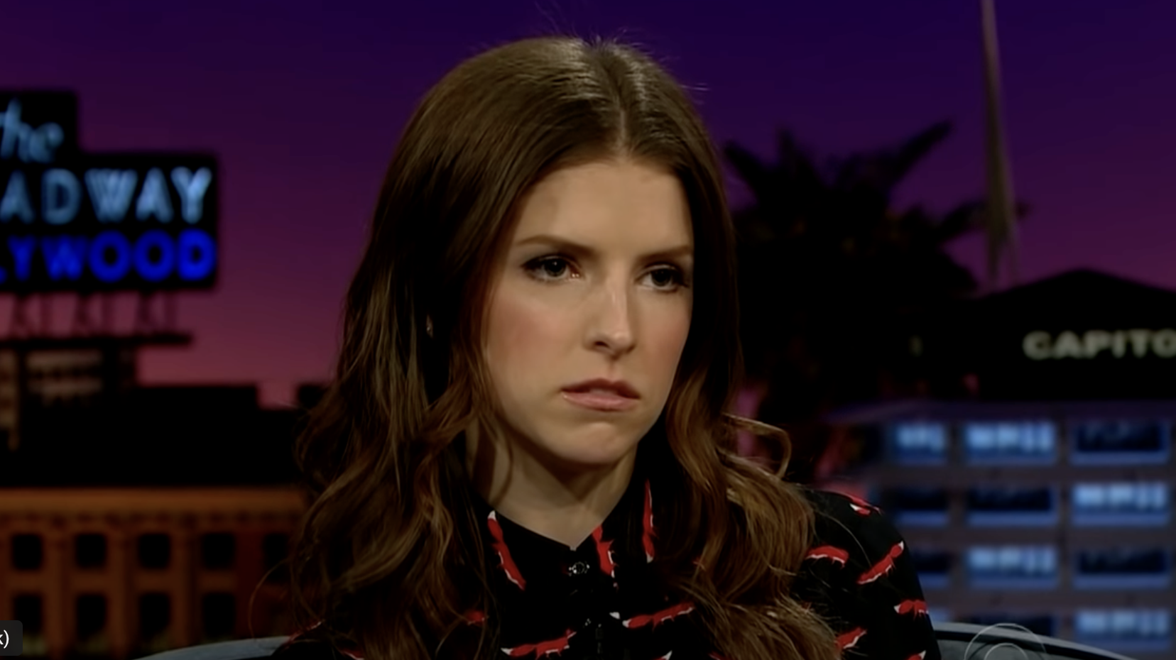 Woman with a concerned expression wearing a printed blouse, sitting in a TV show setting