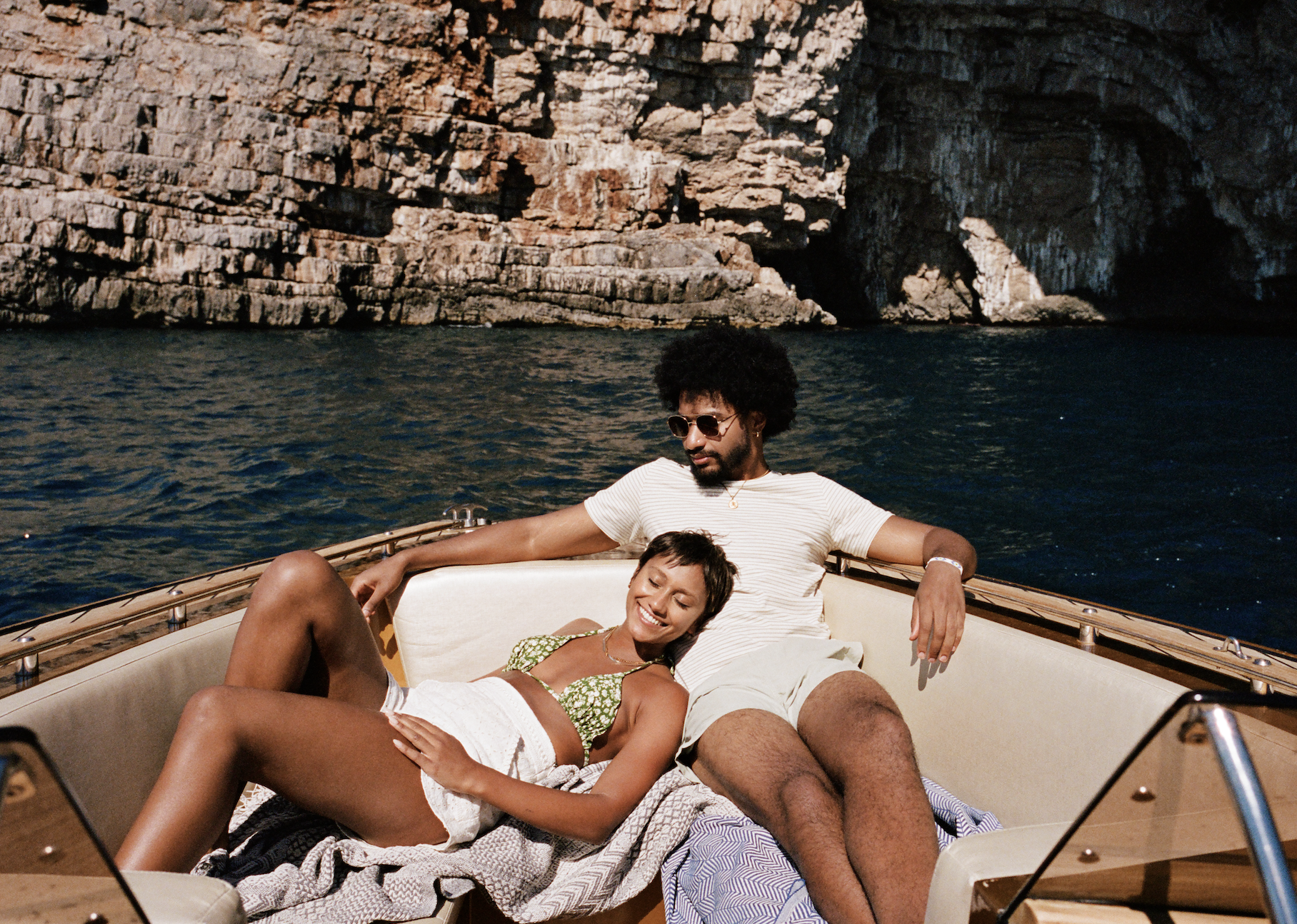 A couple lounges together on a boat with a rocky cliff in the background