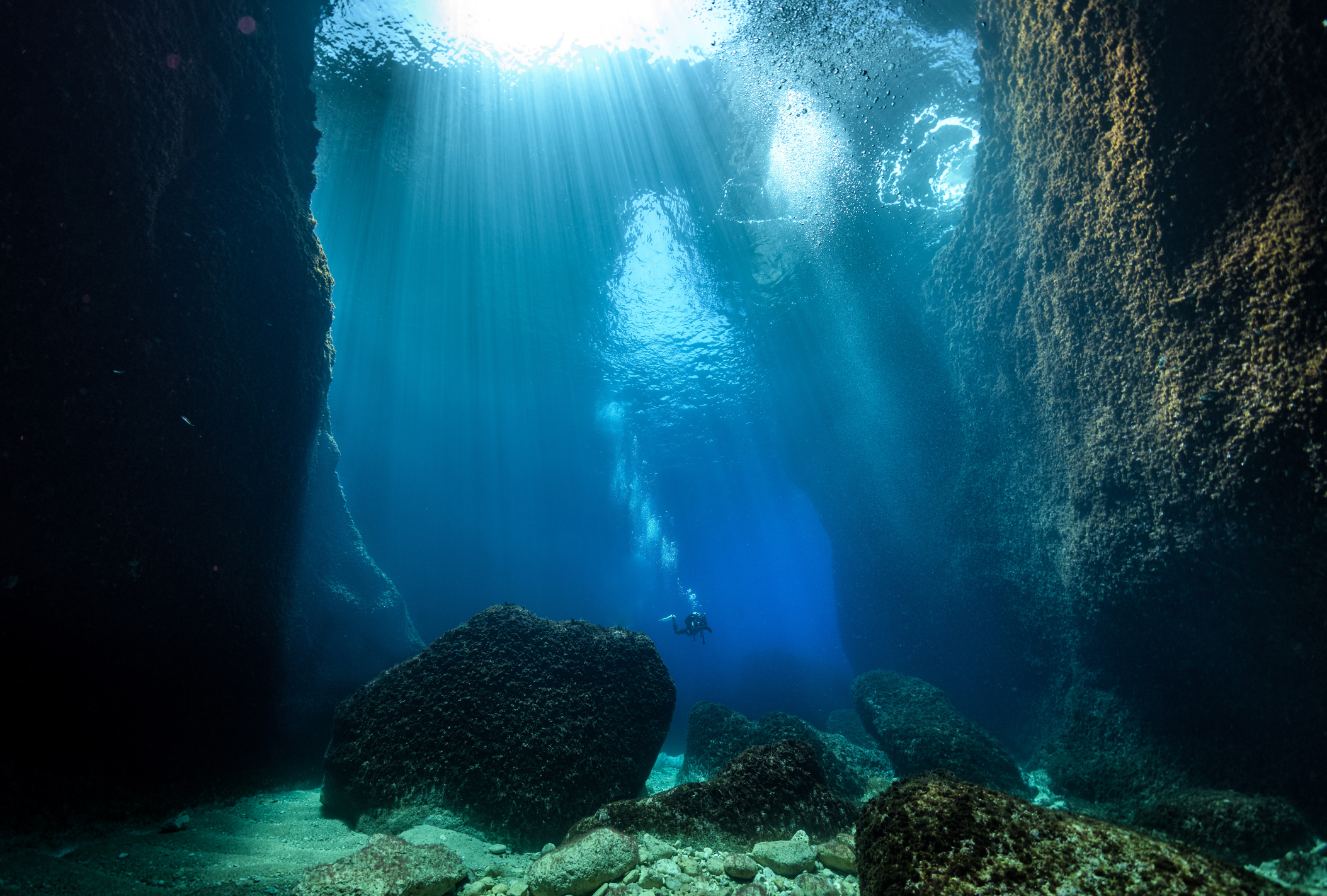 Underwater view of a scuba diver exploring between two large rocks with sunlight piercing through the water from above
