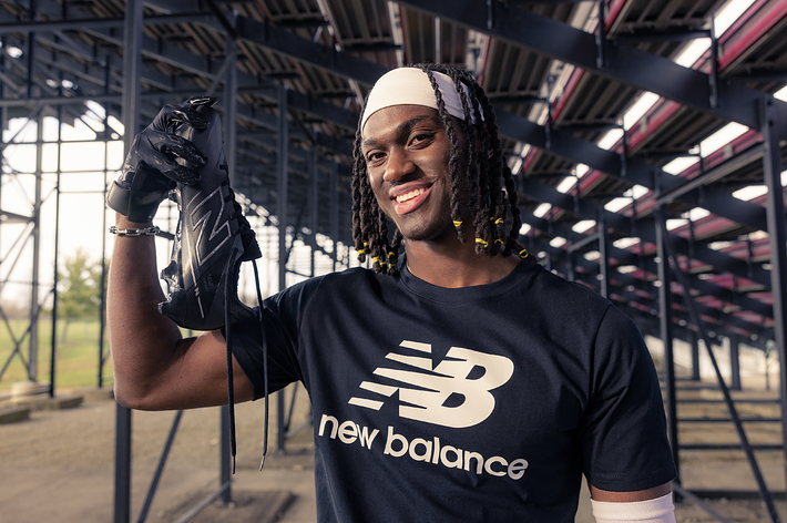 Person smiling, holding a New Balance sneaker, wearing a branded T-shirt, indoors with structural background