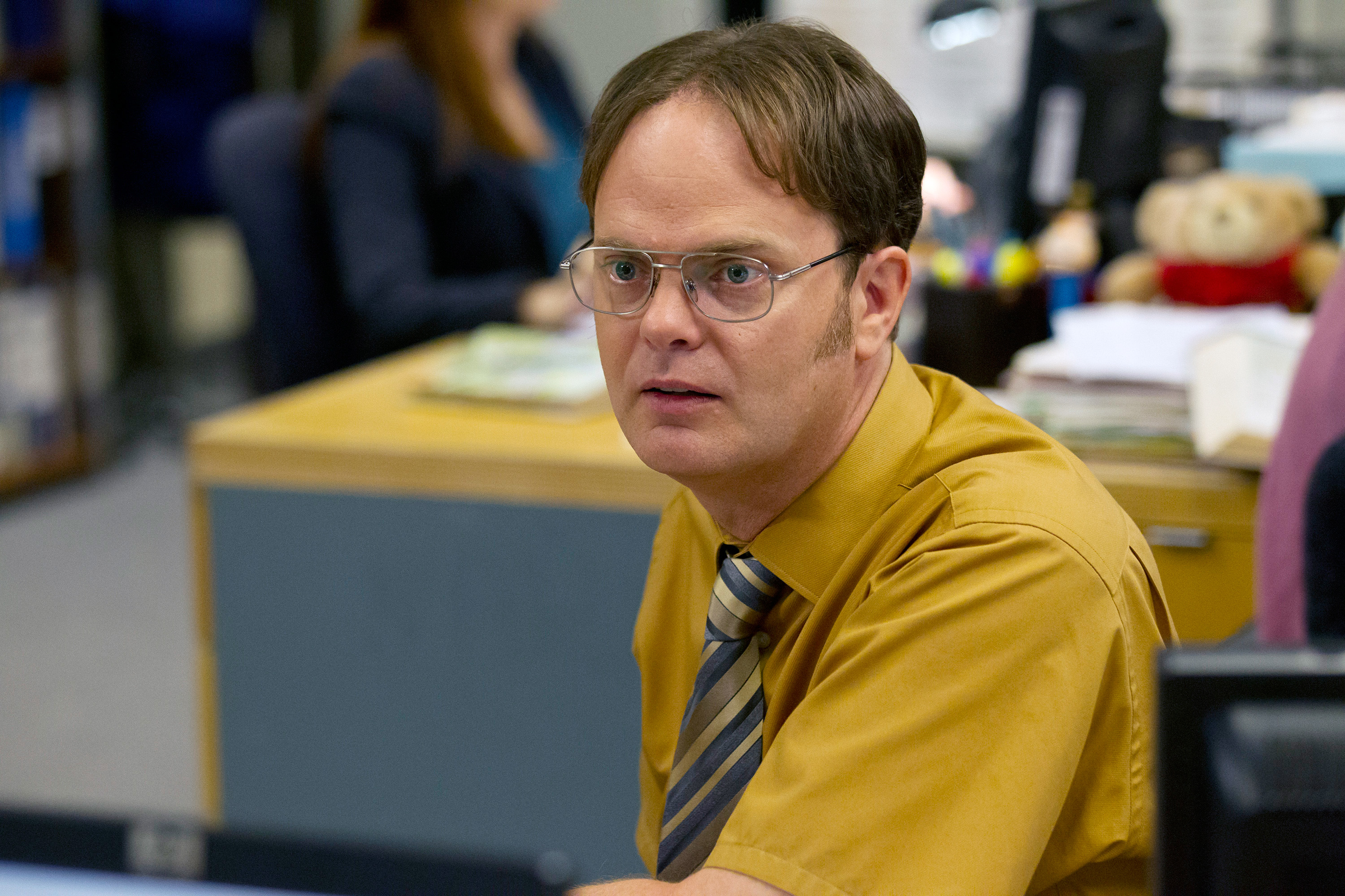 Dwight Schrute from The Office sitting at a desk with a focused expression, wearing a mustard shirt and striped tie