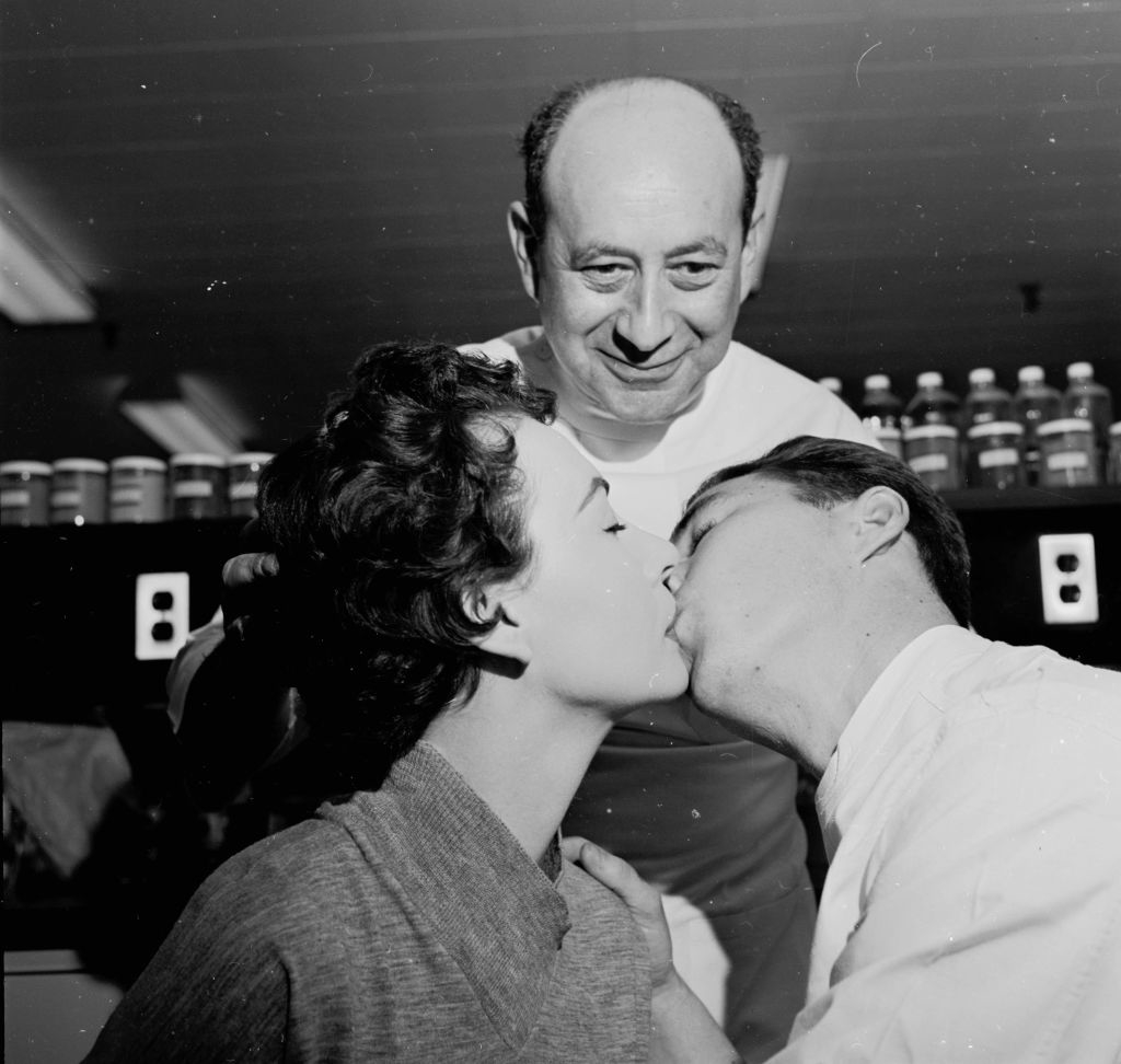 Two individuals sharing a kiss with a smiling person looking on in the background