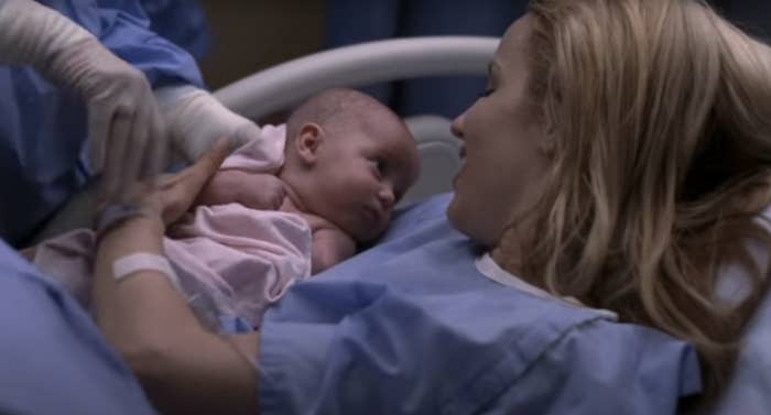 Woman in hospital bed holds a newborn baby, a medical professional assists. (Characters from a TV show)