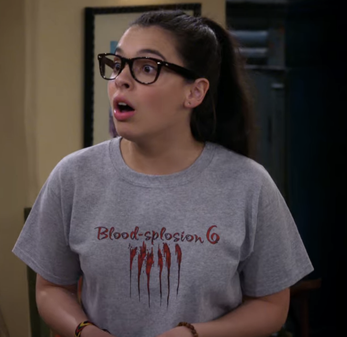 Woman in glasses and a T-shirt with &quot;Blood-splosion 6&quot; text and graphic. She looks surprised