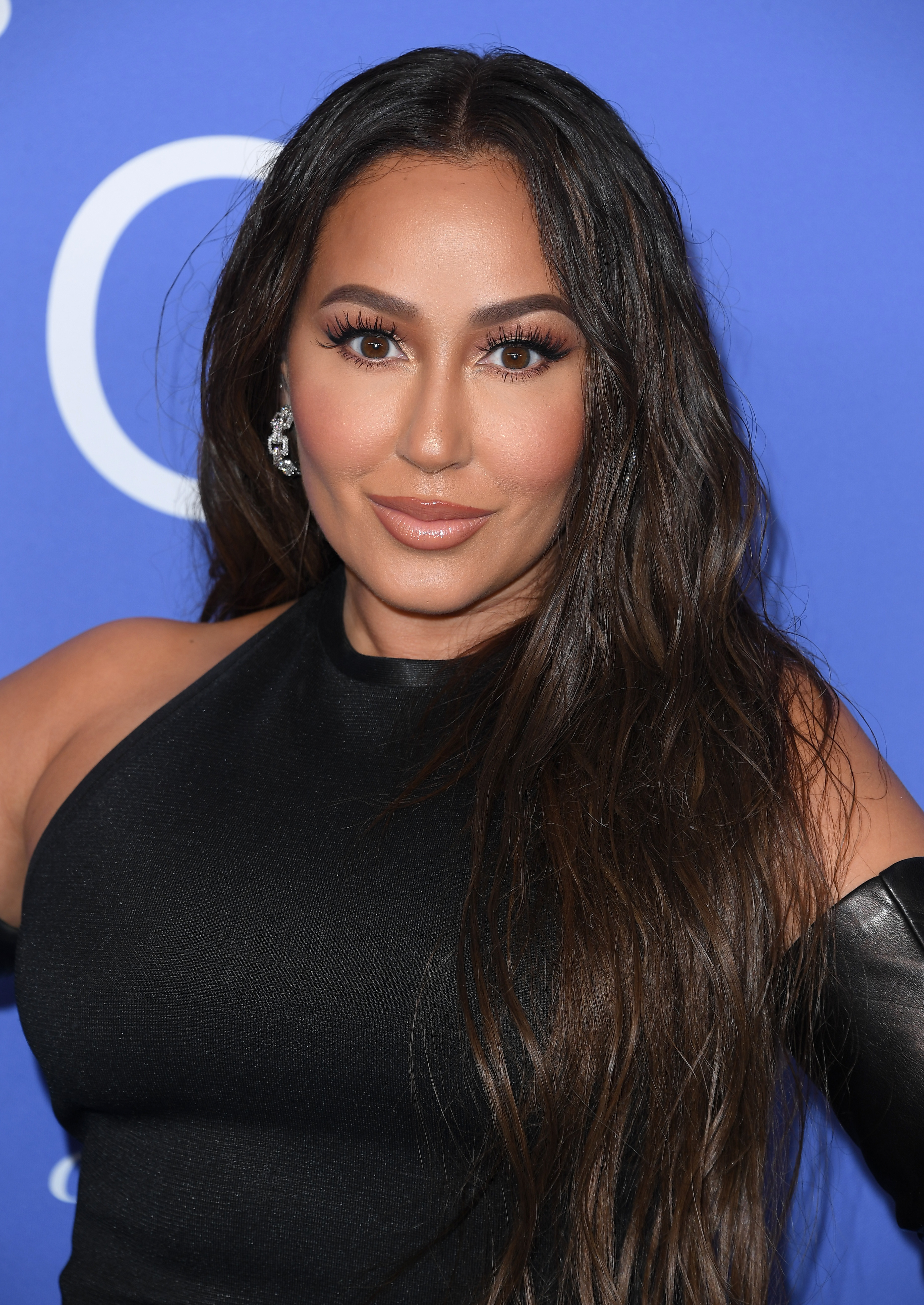 Adrienne Bailon-Houghton wearing a black one-shoulder top, posing for the camera