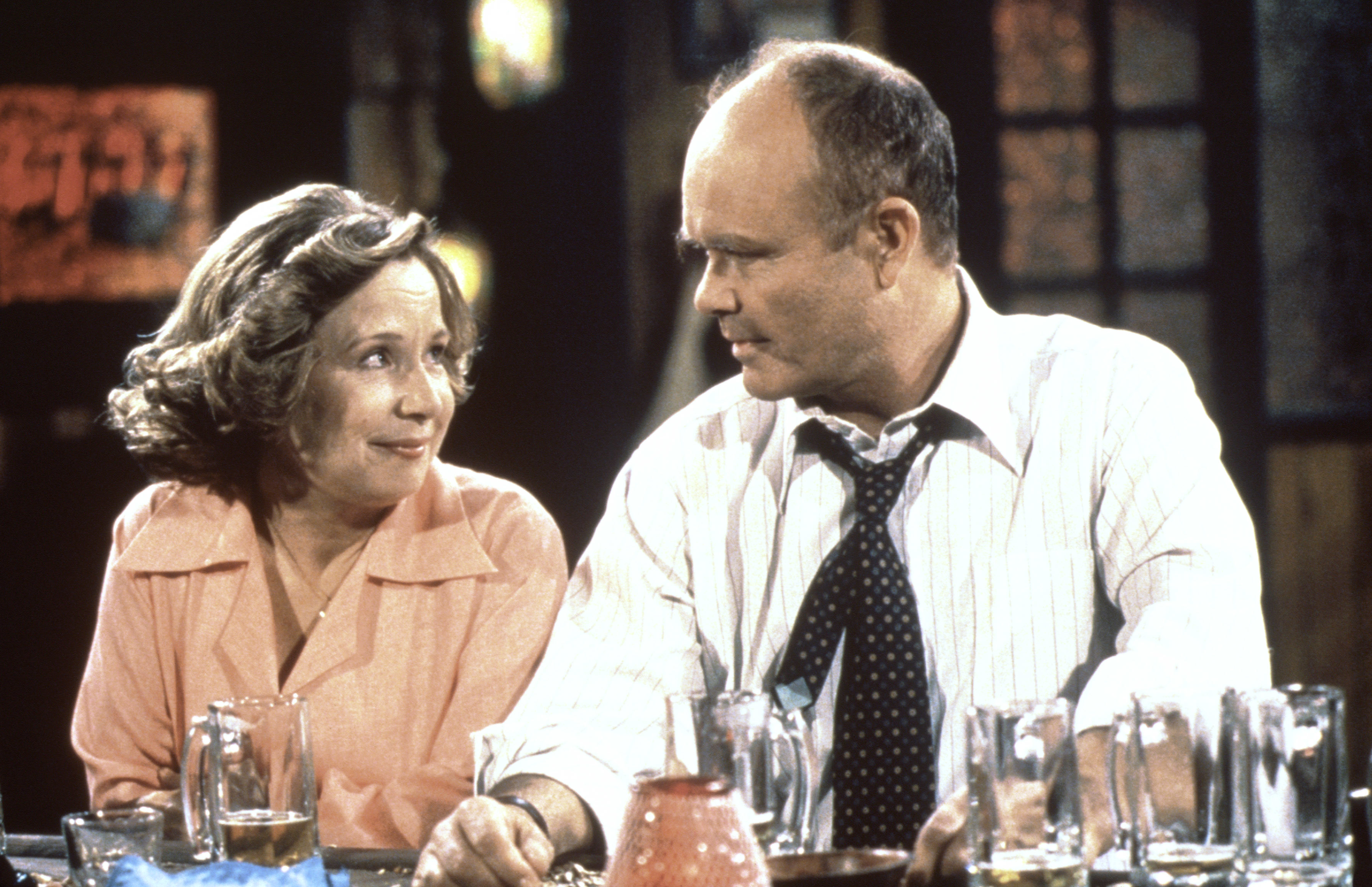 Two actors on set, sitting at a table with glasses, portraying characters in a conversation