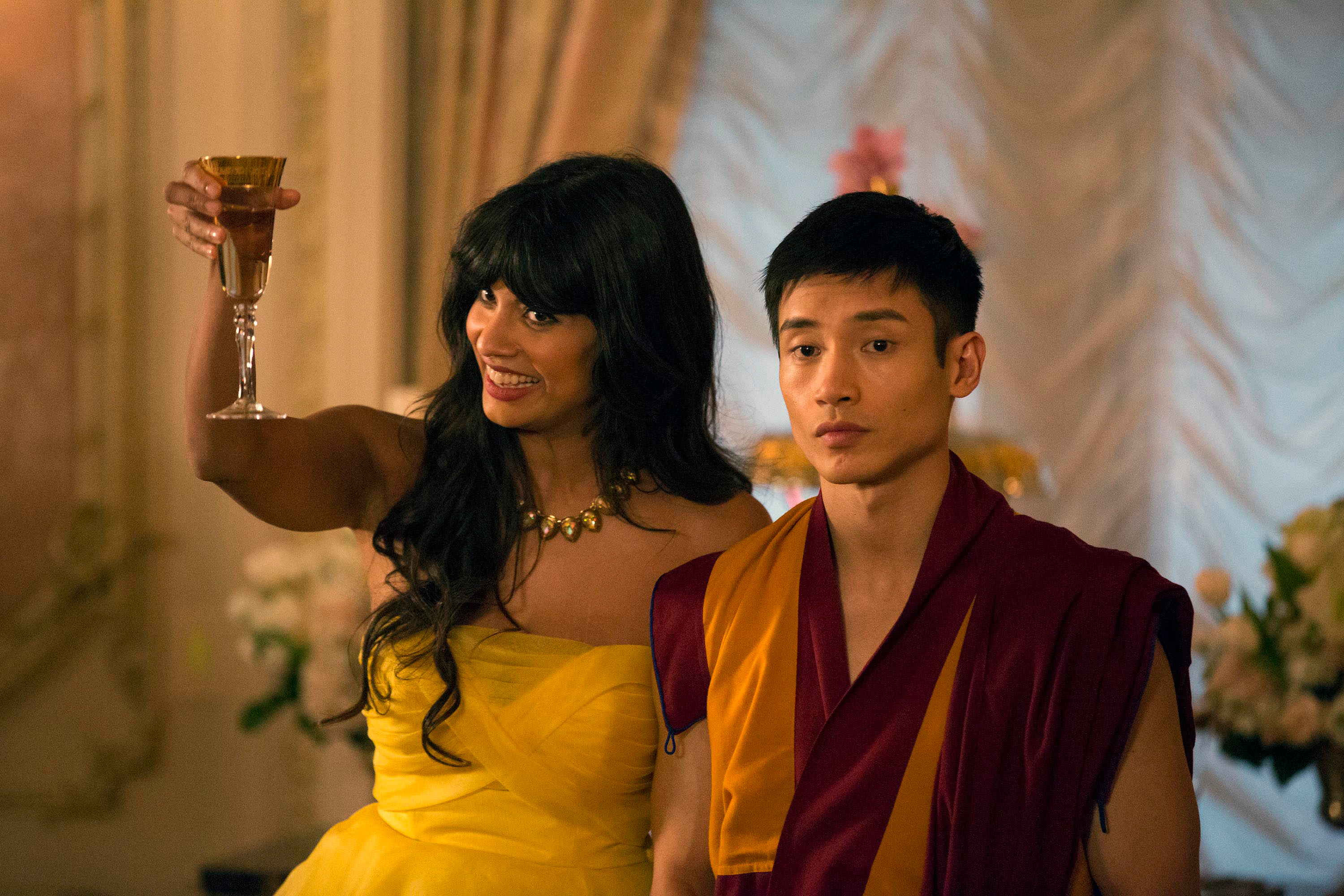 Two characters from TV show, woman in yellow dress with a glass, man in monk robes, indoors