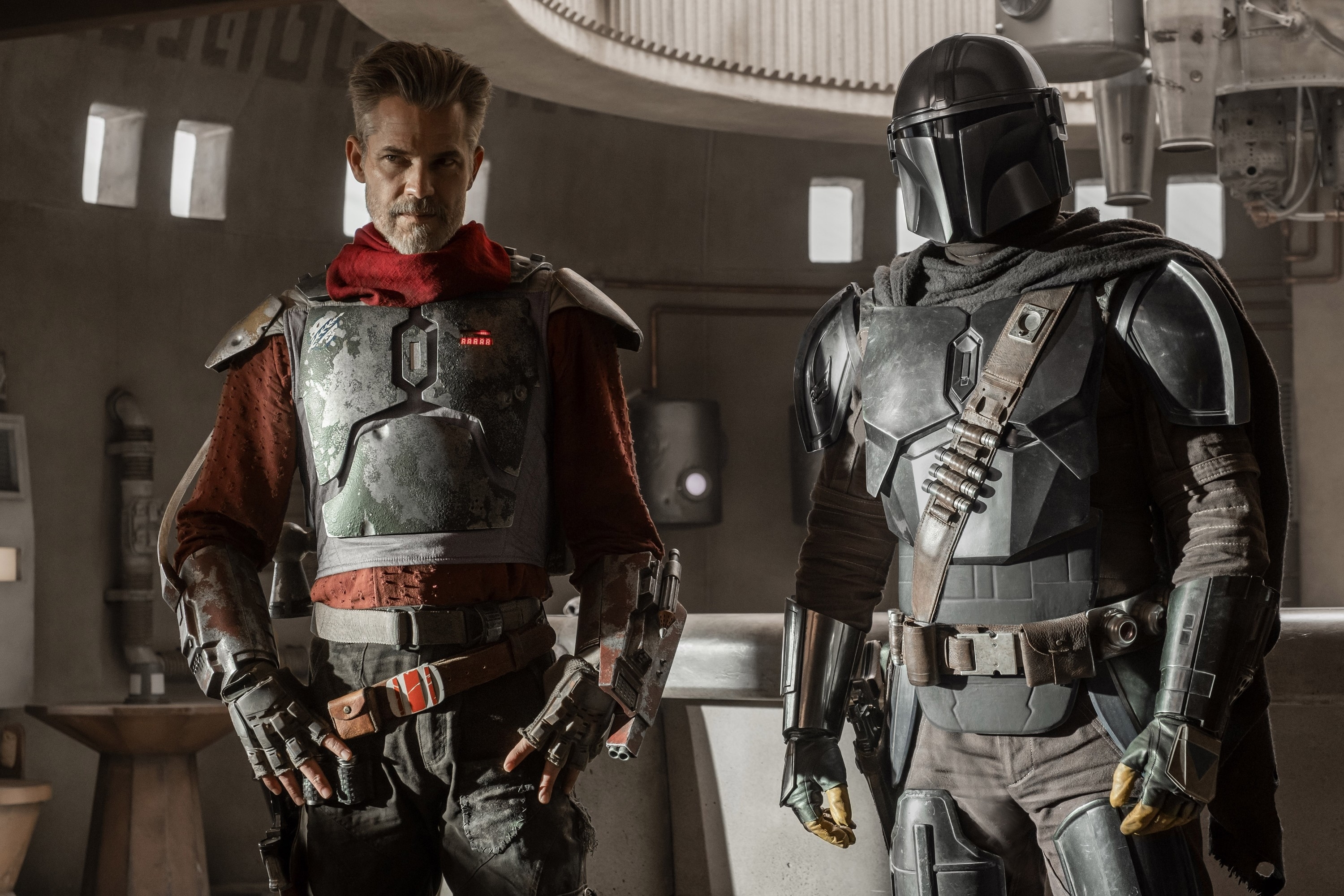 Two characters from The Mandalorian, one in a red suit with armor and the other in a silver armored suit, stand side by side