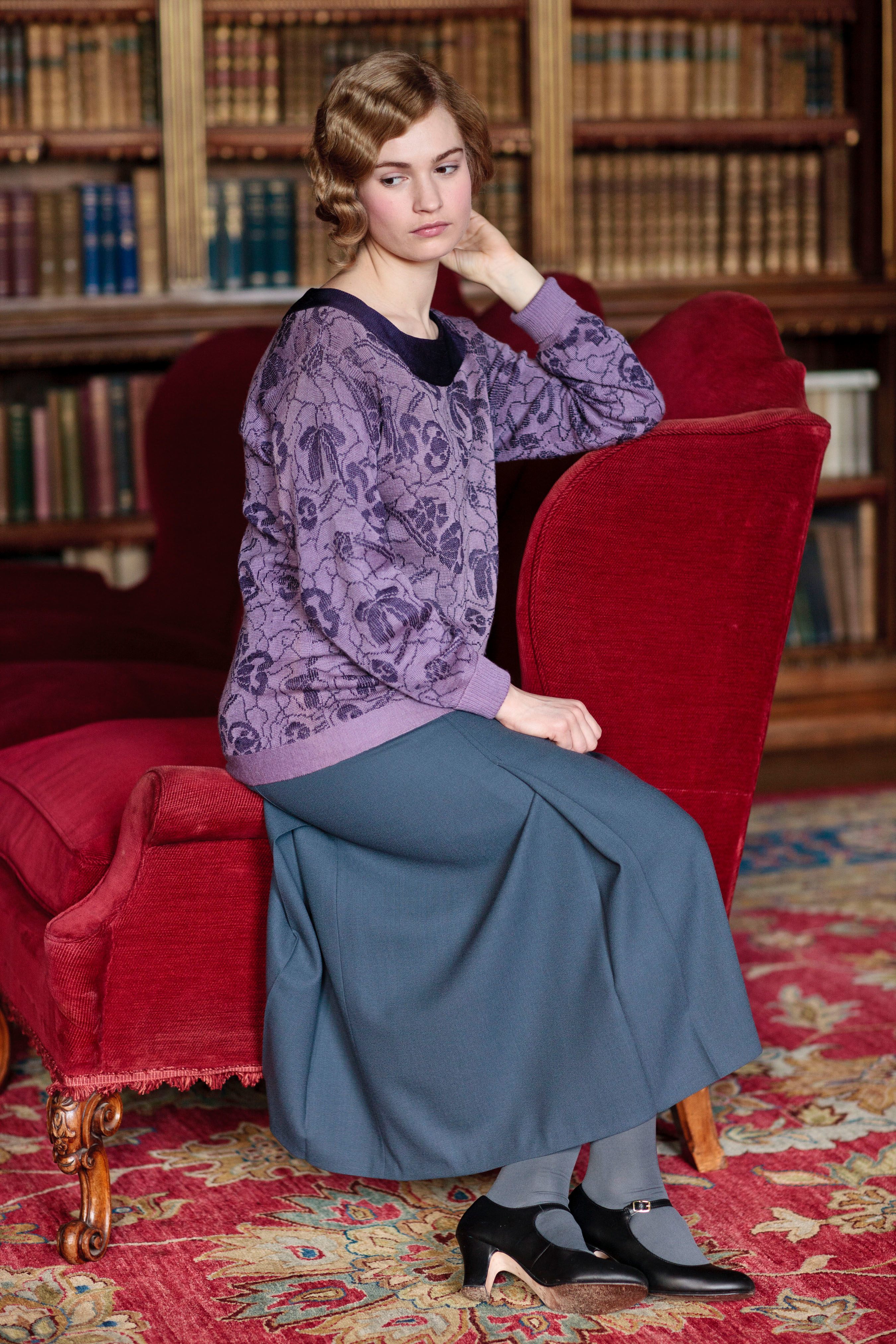 Woman posing in a vintage style purple blouse and grey skirt, seated on a red chair in a library