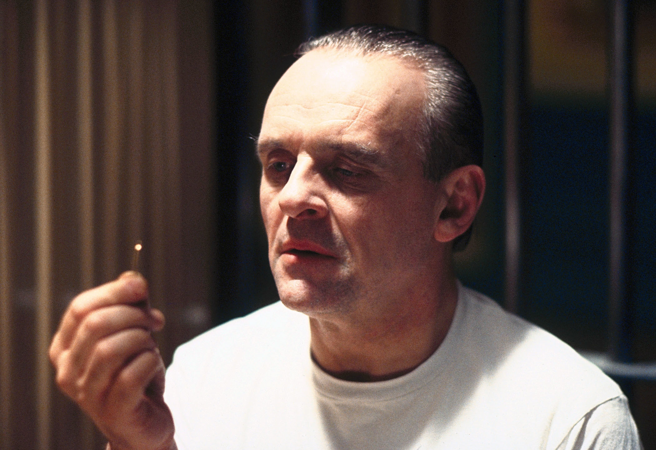 Man in white T-shirt sitting, holding a toothpick, with a focused expression