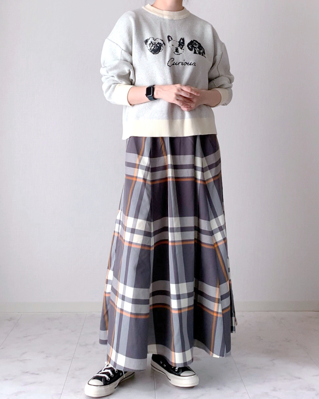 Person in a grey sweatshirt and plaid skirt standing with arms crossed, wearing sneakers