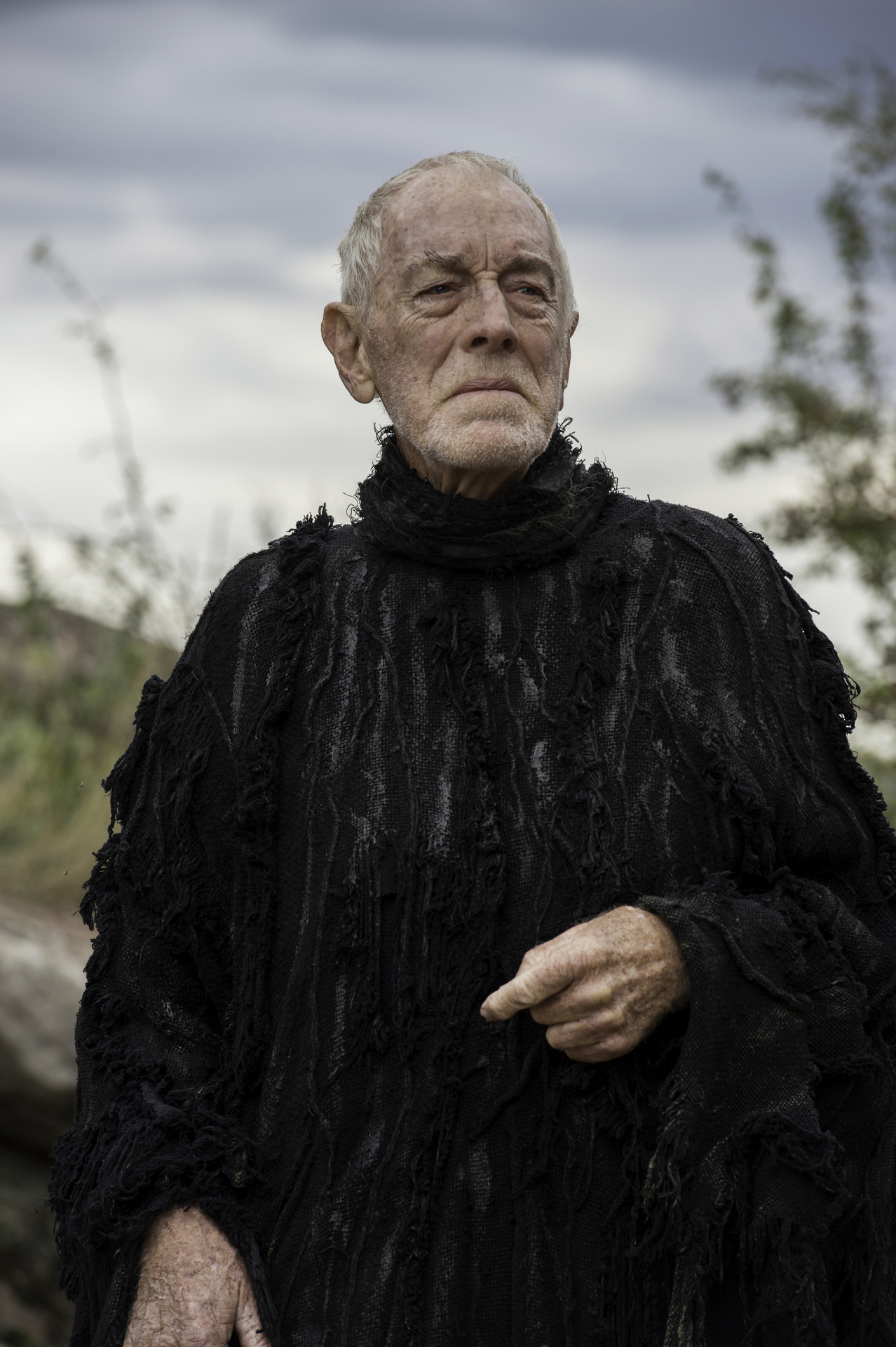 An older man in a textured black outfit stands outdoors, looking deeply into the distance
