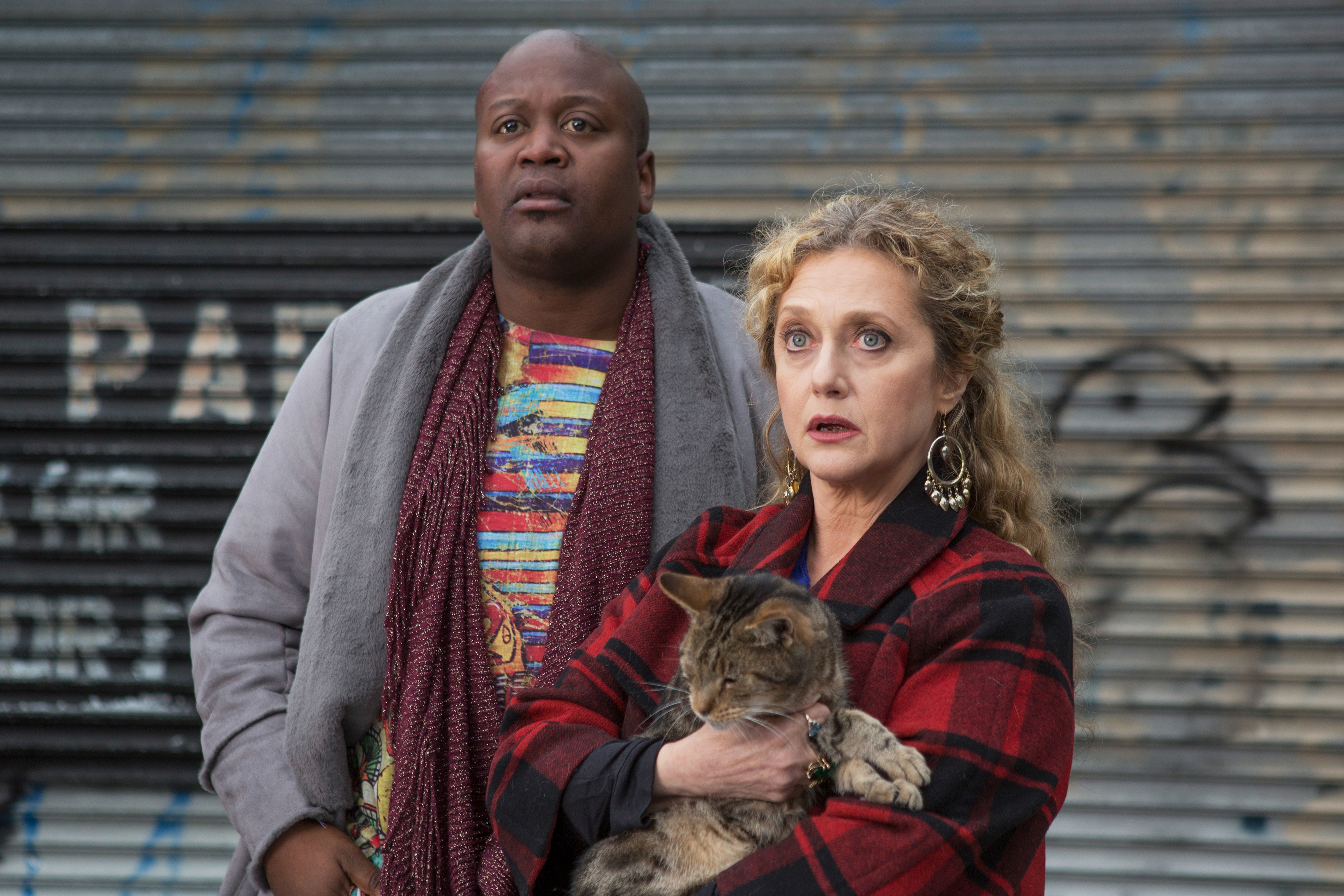 Two actors from the TV show &#x27;Kimmy Schmidt&#x27; posing with a cat, man in scarf and woman in plaid coat