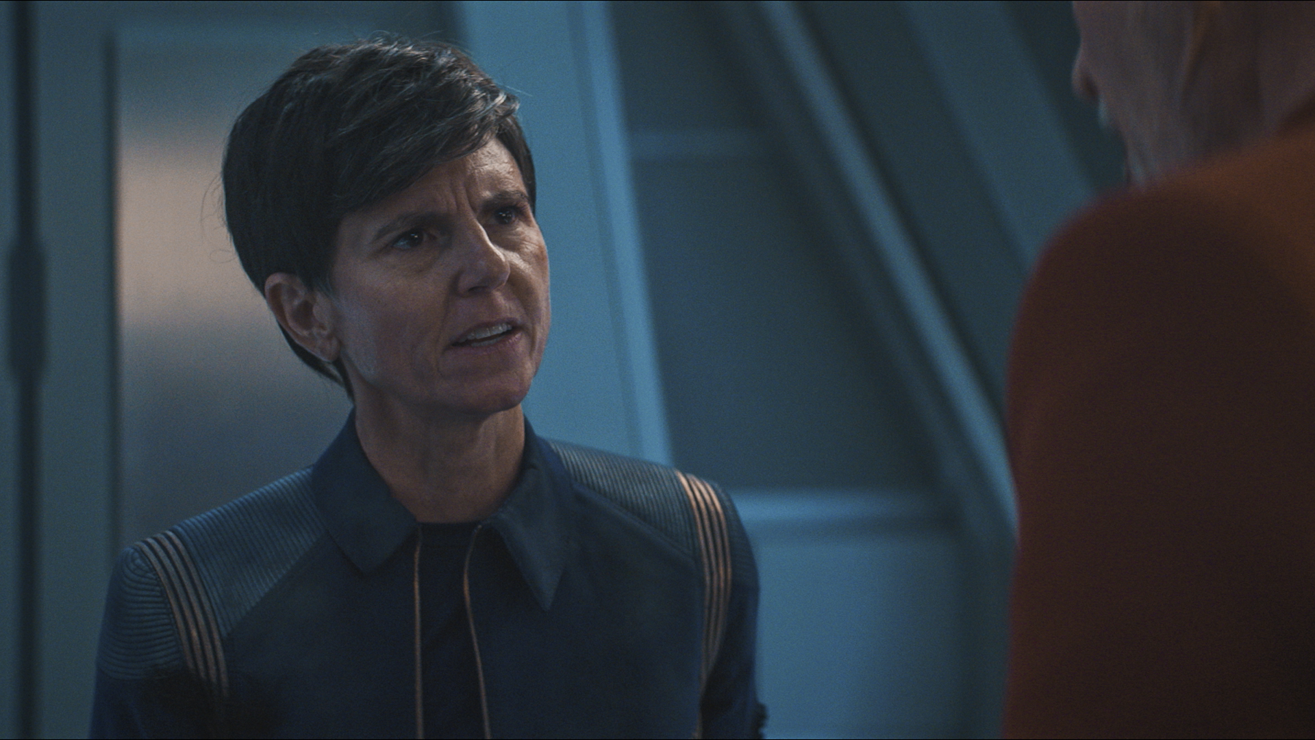 Tig Notaro, dressed in a blue uniform, engaged in a serious conversation on a spaceship set