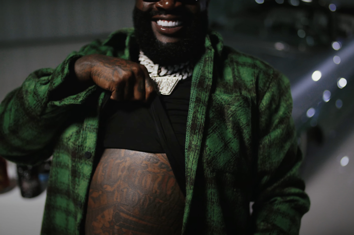 Person in black outfit with a green jacket, showing tattooed arm and neck jewelry