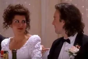 Elaine in a white poofy dress and Jerry in a tuxedo sitting at a table, looking amused