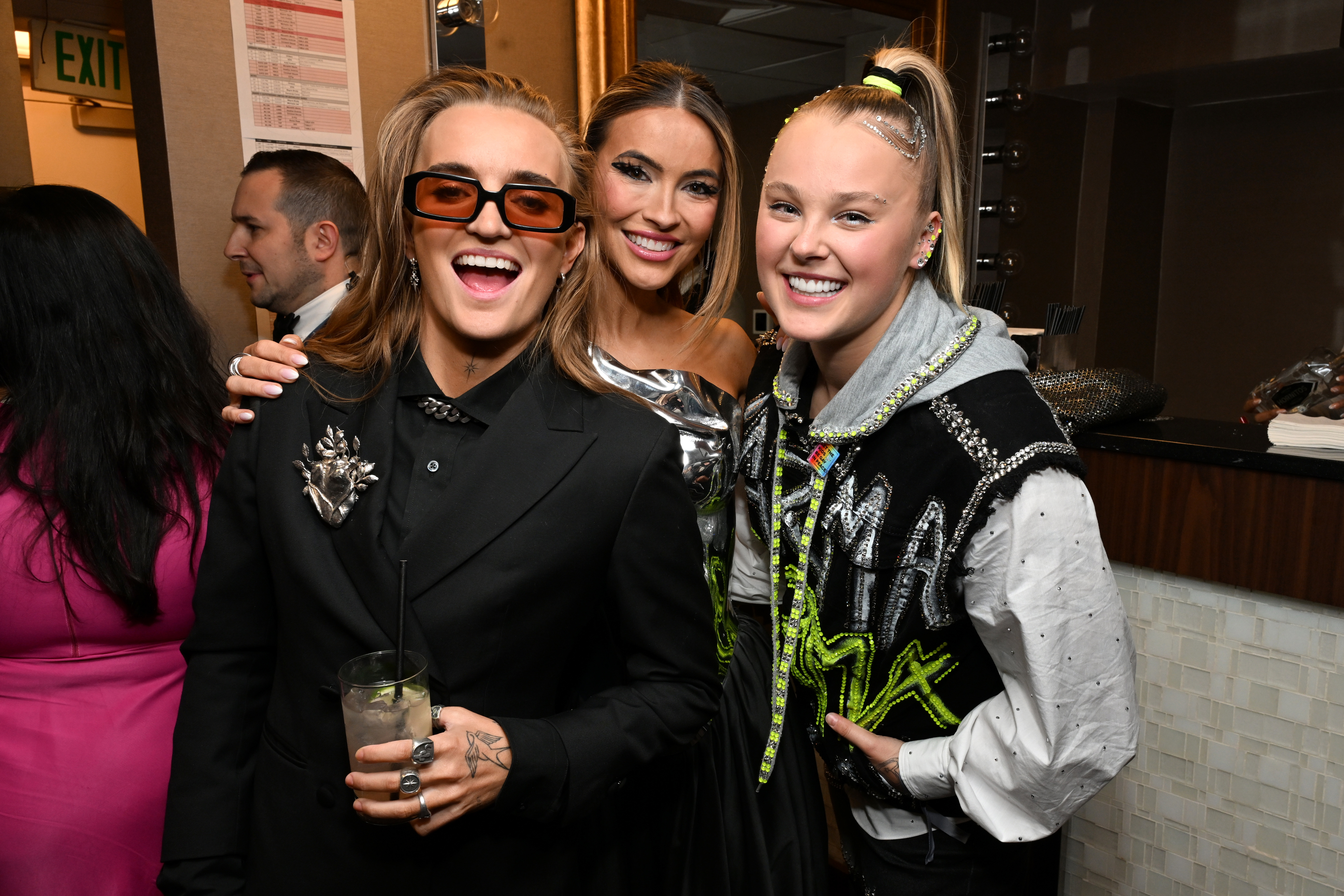 Three smiling individuals pose together at an event, two in embellished black jackets and one in a metallic dress