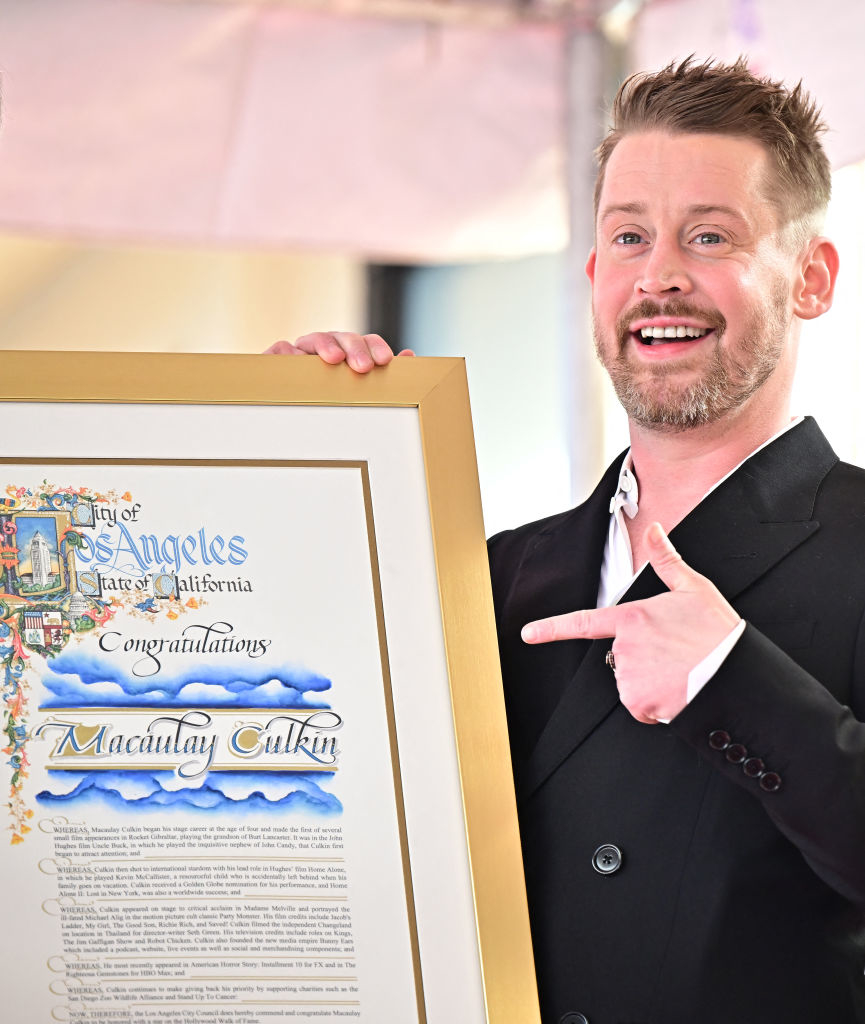 Man in suit holding a framed certificate with the name &quot;Macaulay Culkin&quot; on it, smiling and pointing at the text