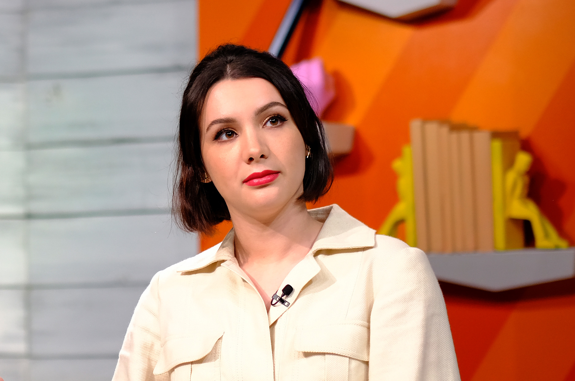 Hannah Marks in a beige jacket, on a set with various decor in the background