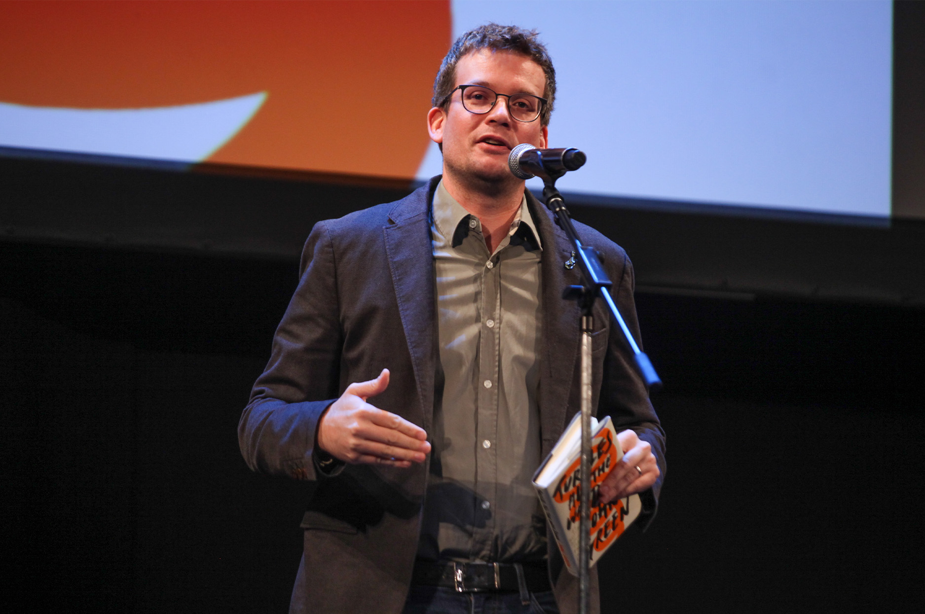 John Green in a blazer and glasses speaking at a microphone with a book in hand