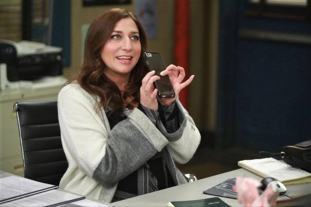 Woman seated at a desk smiling and holding up a cell phone, portraying the character Gina Linetti from Brooklyn Nine-Nine