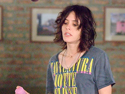 Woman in a graphic t-shirt and disheveled hair holding a bottle. She portrays a character in a casual indoor setting