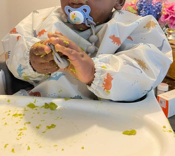 a reviewer photo of a baby with a pacifier wearing a bib, hands smeared with food at a high chair