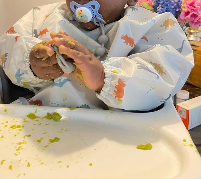 a reviewer's baby with a pacifier wearing a bib, hands smeared with food at a high chair