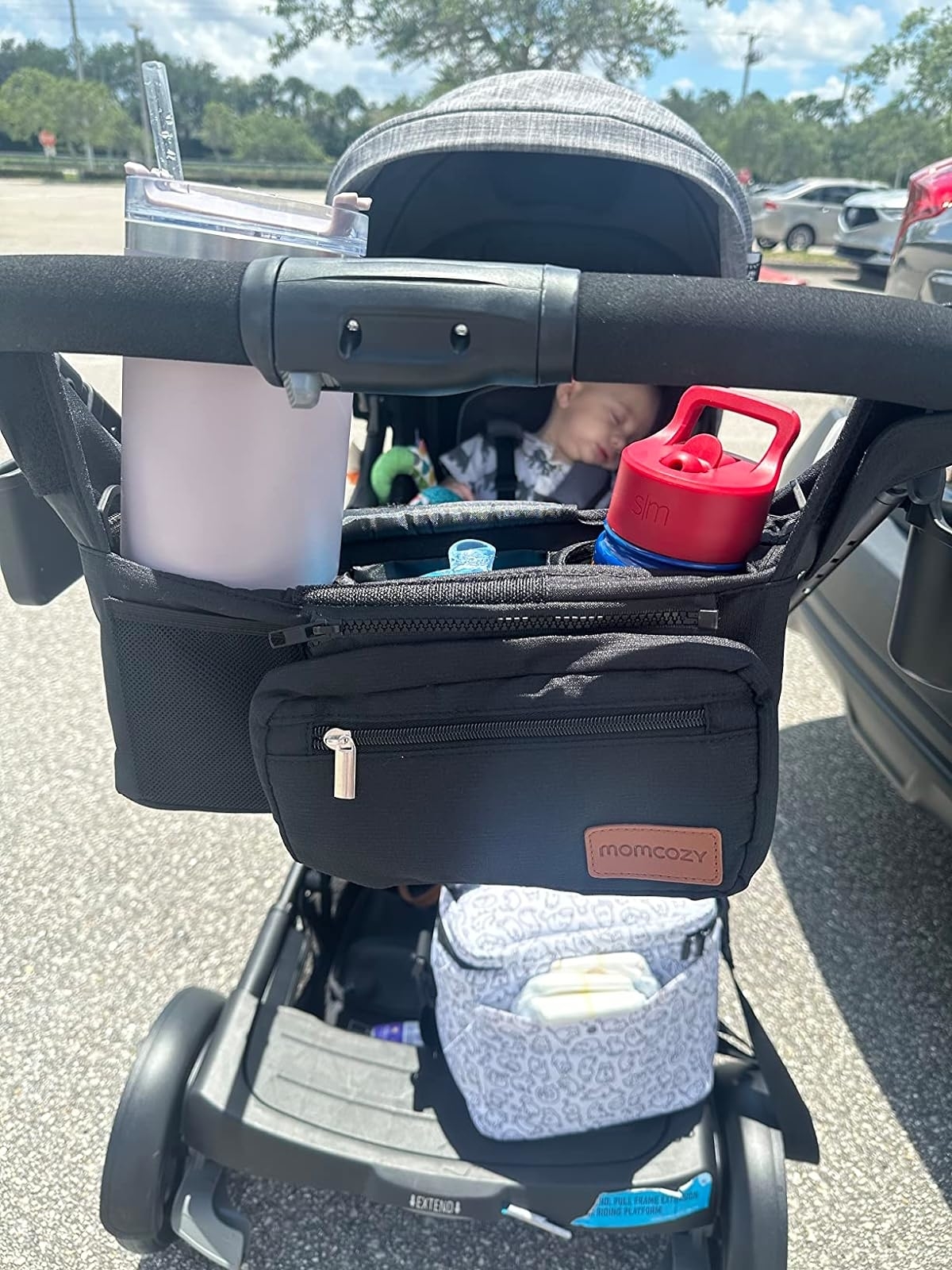 Baby asleep in a stroller with an organizer holding a bottle and a drink. A bag and a burp cloth are underneath