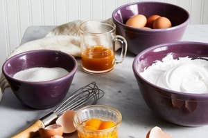 Purple mixing bowls in three different sizes filled with eggs, flour, sugar, and liquid, whisk and cracked eggshells visible on counter