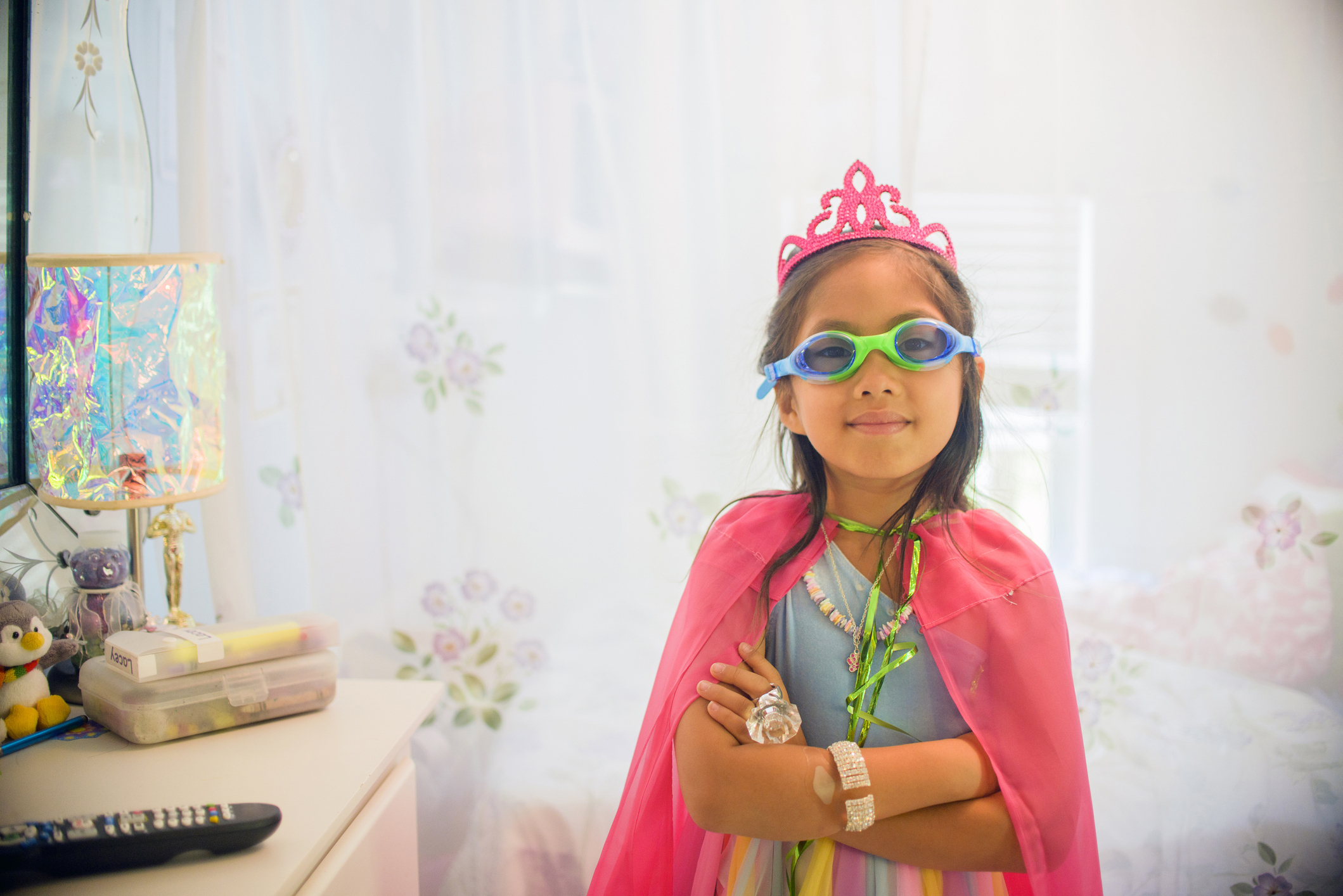 Child in a cape and tiara with goggles, posing confidently in a bedroom