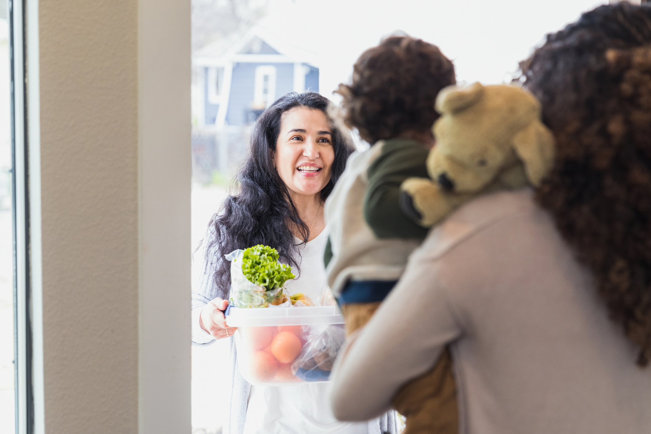 Woman holding a food bowl smiling at another woman carrying a child with a plush toy. They are indoors, possibly at a gathering