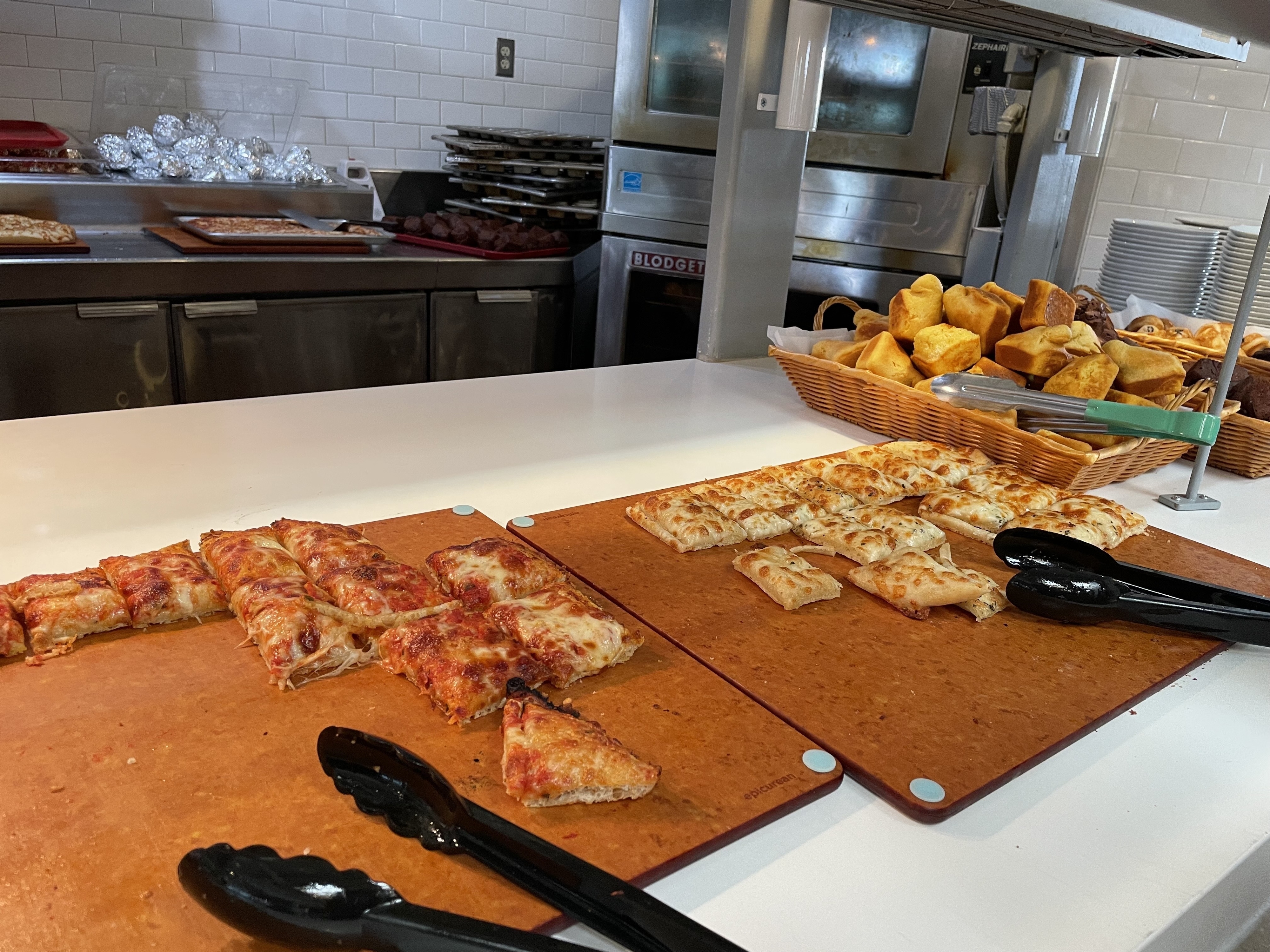 Assorted slices of pizza on a wooden board with tongs, next to a basket of garlic knots in a kitchen setting