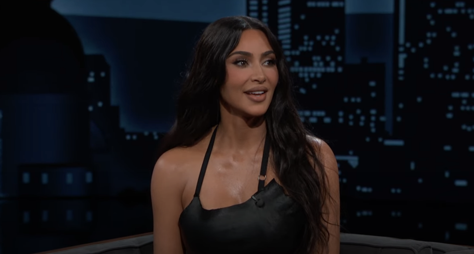 Kim Kardashian in a talk show setting, wearing a strappy top, seated with a cityscape backdrop