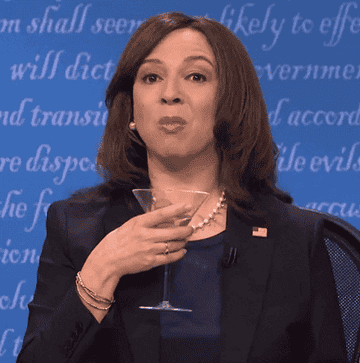 Maya Rudolph impersonating Kamala Harris with a beverage, expressing with hand gestures, wearing a suit and pearls