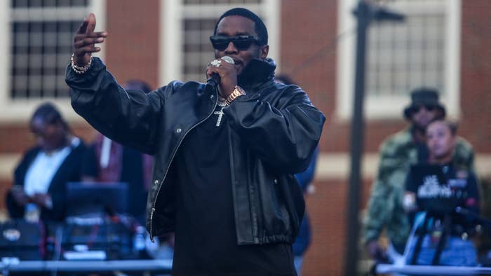 Diddy on stage performing with microphone, wearing a jacket and sunglasses, with DJ in background