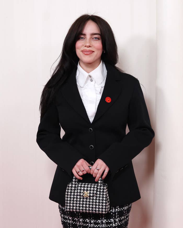 Billie Eilish standing with a black blazer, white shirt, handbag, and sequined bottom