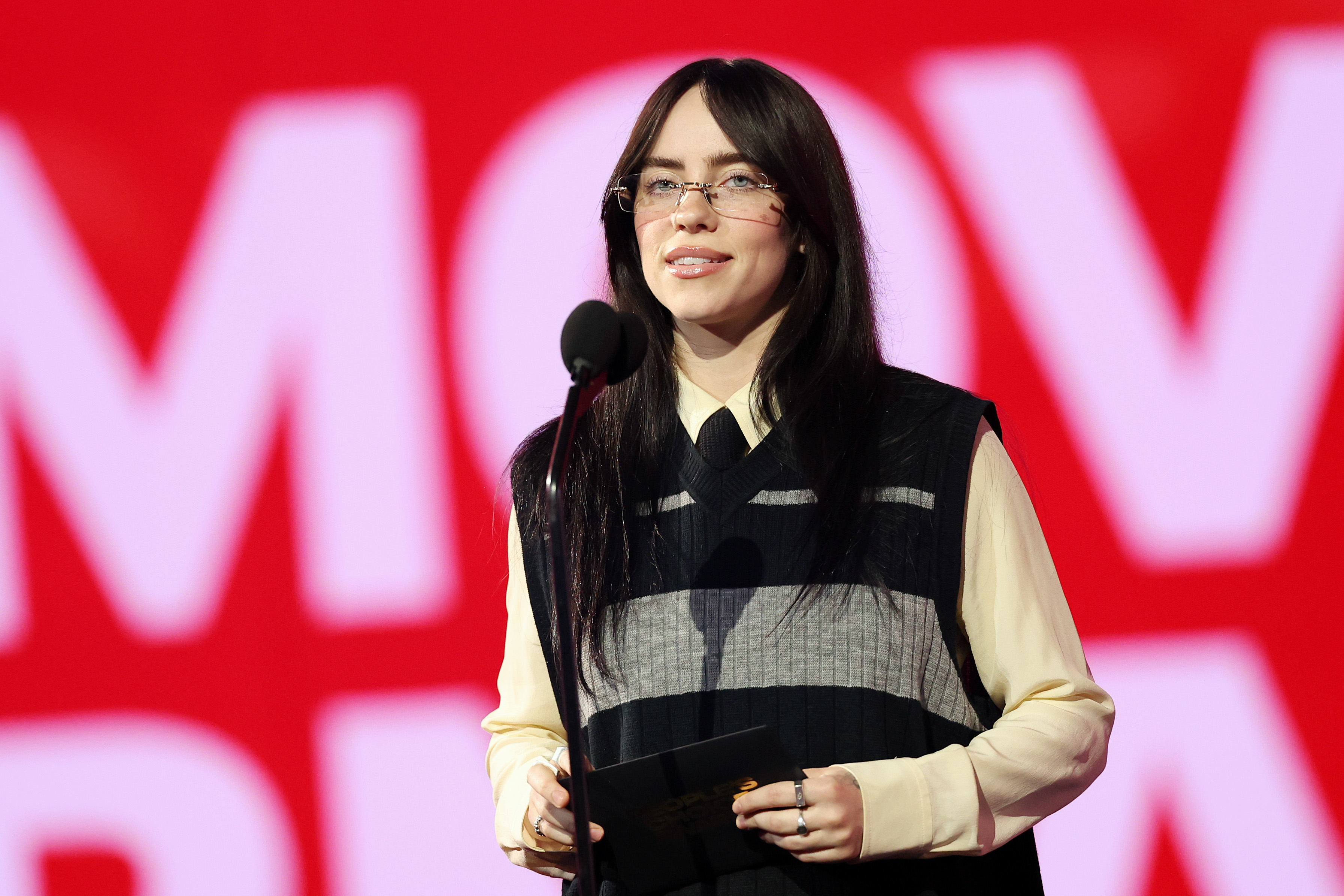 Billie Eilish stands at a microphone wearing a sleeveless sweater over a shirt
