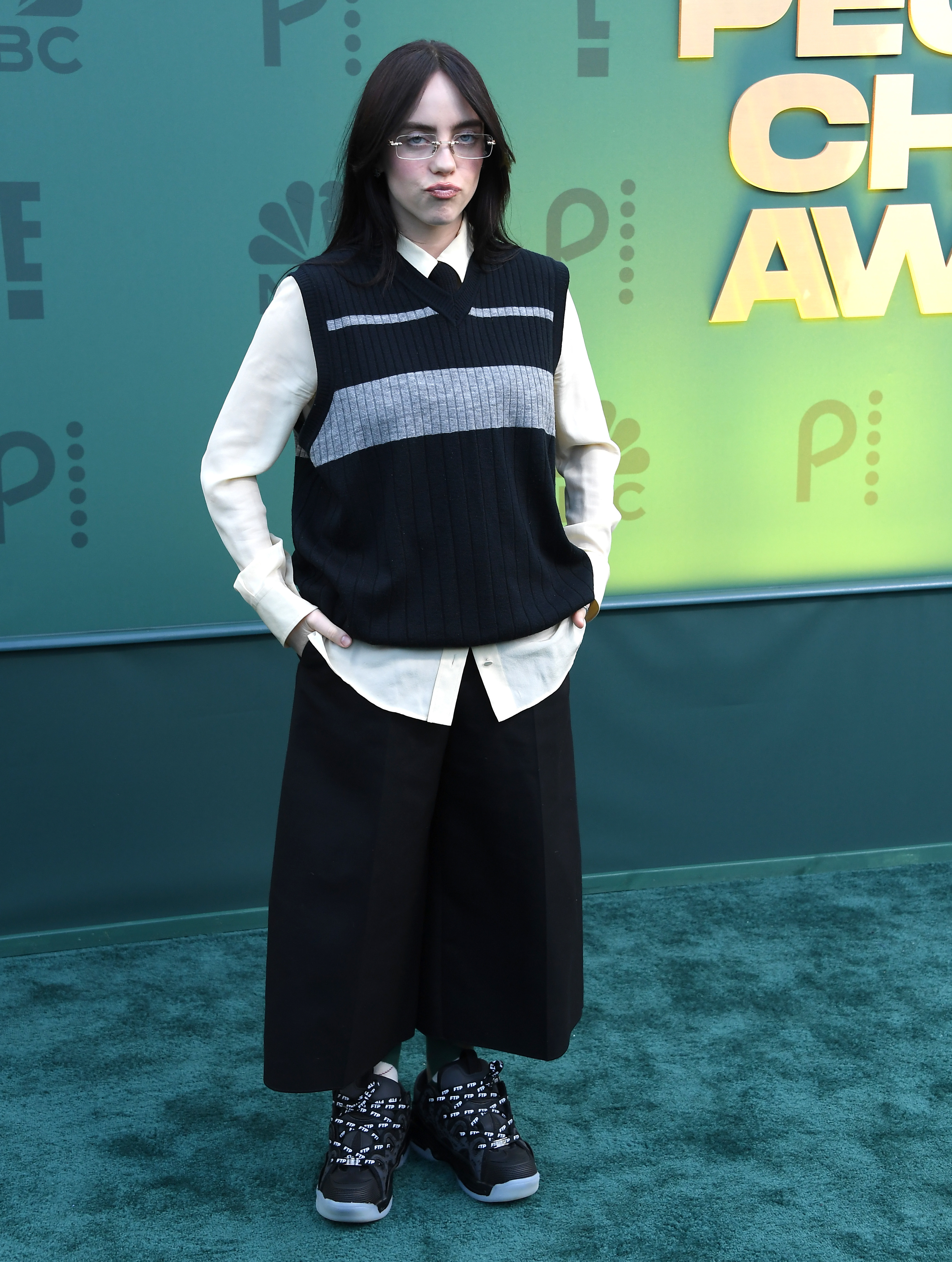 Billie Eilish standing with hands in pockets, wearing a layered oversized outfit and sneakers at an award event