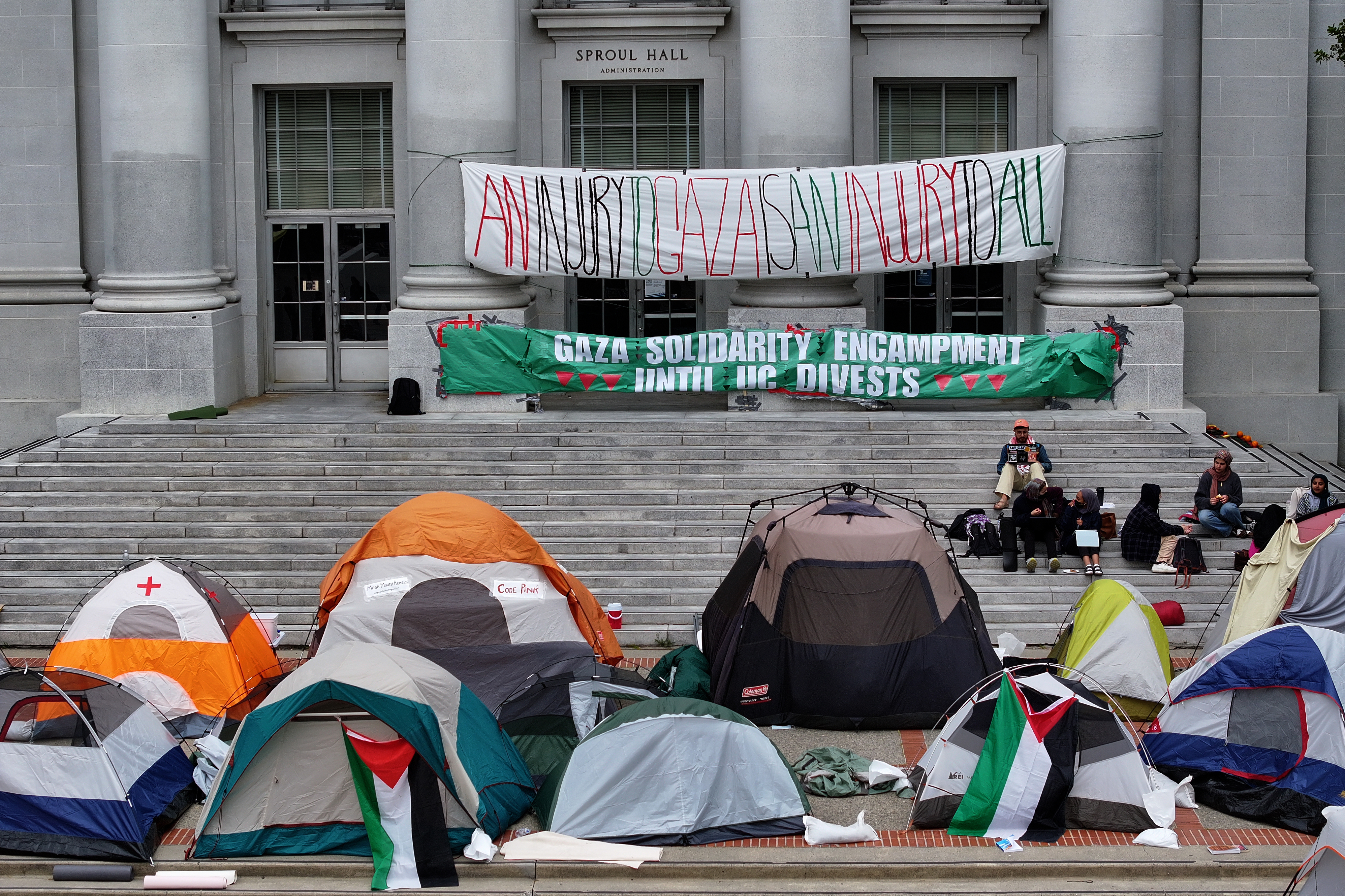 Tents are set up on steps of a building with banners about solidarity and divestment. People are seated around casually