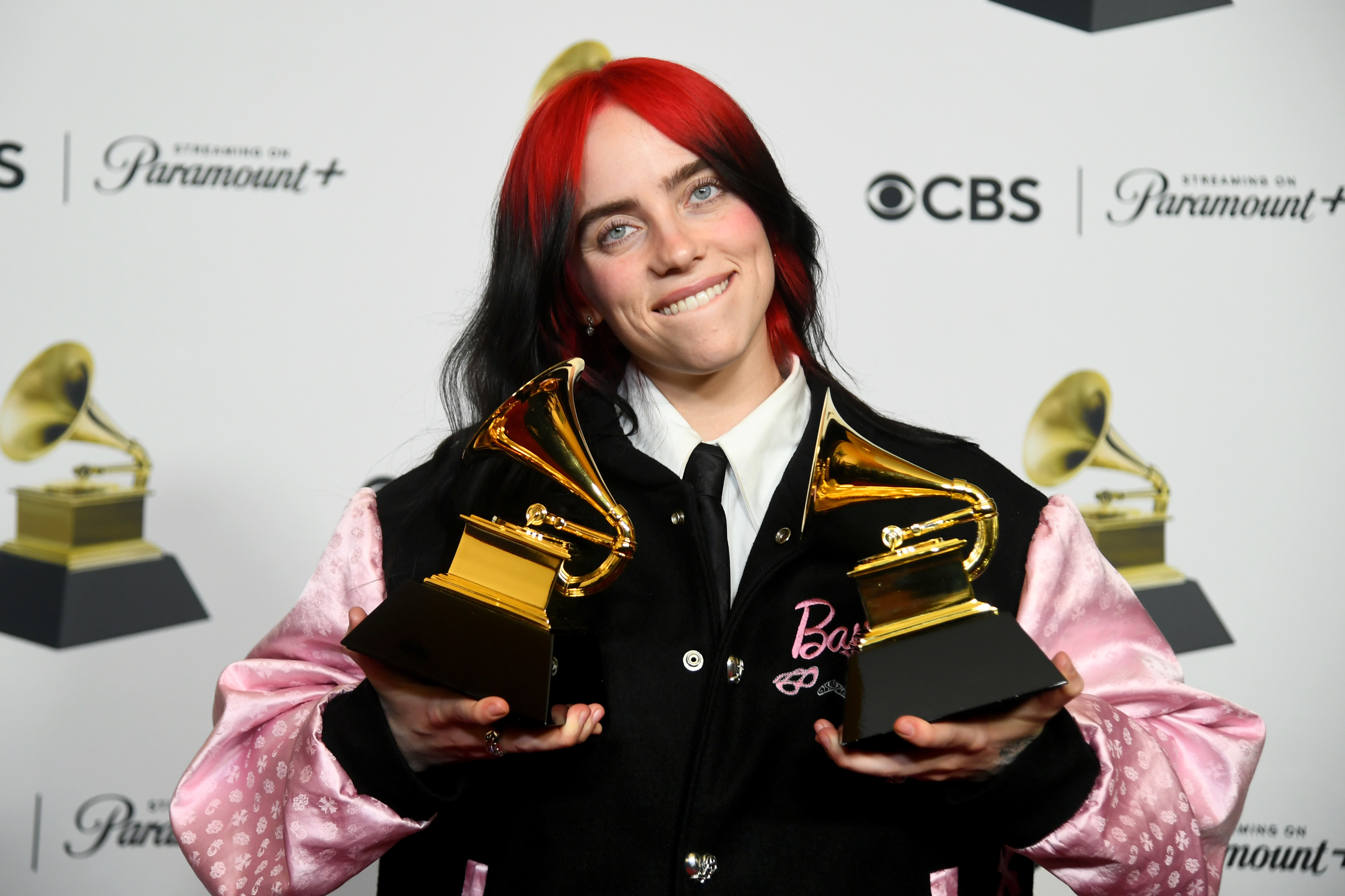 Billie Eilish smiles holding two Grammy awards, wearing a black and pink outfit with the word &quot;Billie&quot; on it