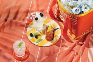 Drinks on a patterned plate with a cooler filled with ice and canned beverages nearby, suggesting a summer picnic setup