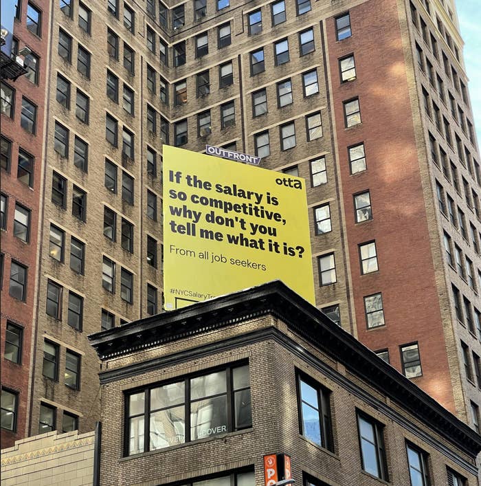 Billboard above city street questioning why competitive salaries aren&#x27;t disclosed, engaging job seekers. #NYCSalary #InDoors
