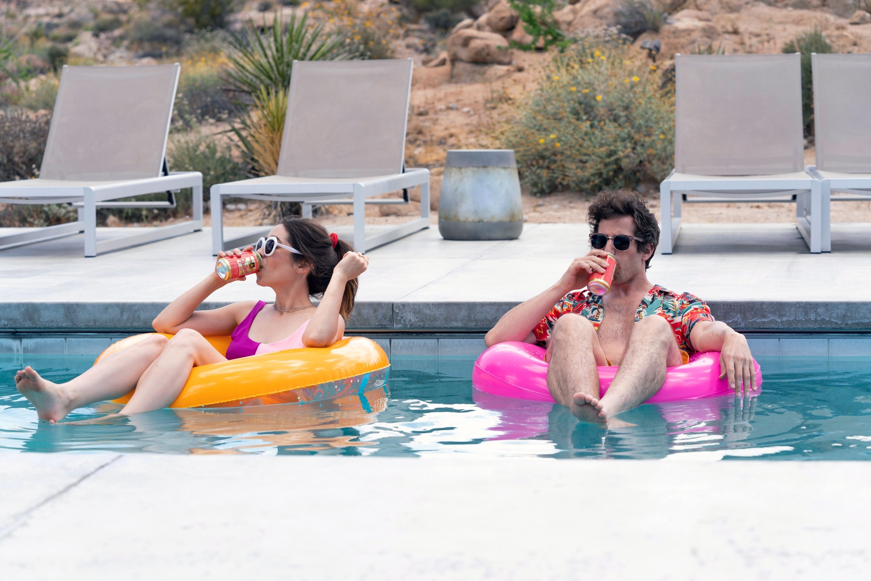 Two people relaxing on pool floats, sipping drinks, in a pool by lounge chairs