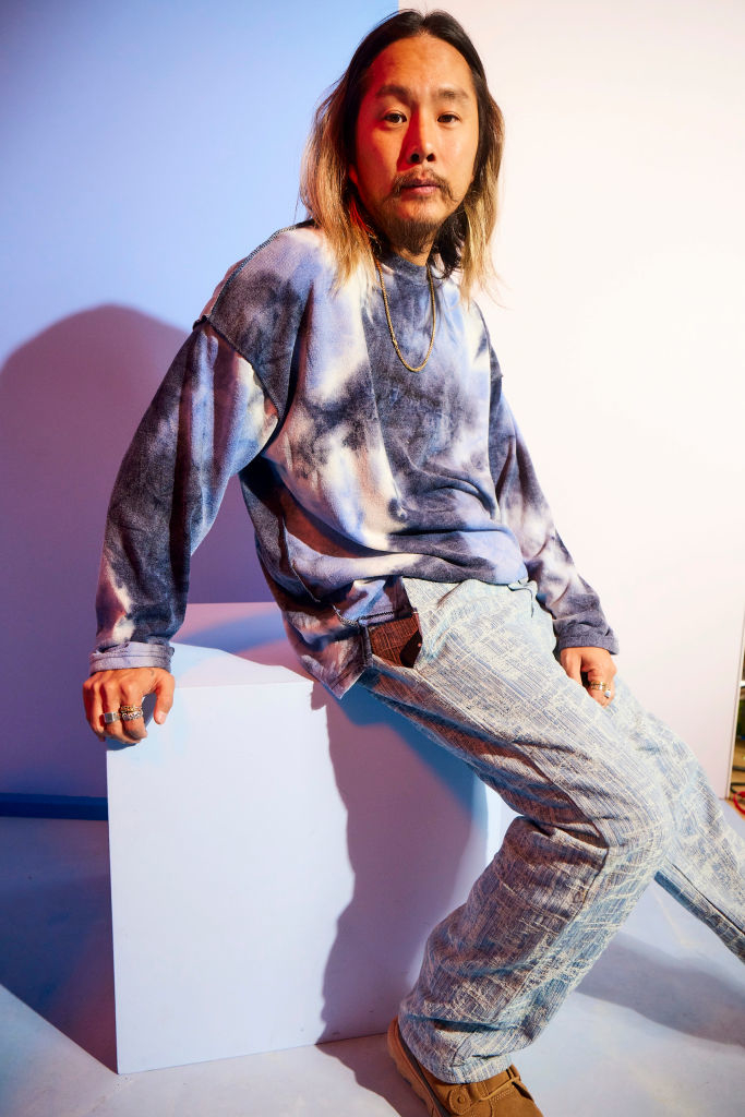 Justin in tie-dye sweatshirt and jeans sitting on a cube, posing with a serious expression