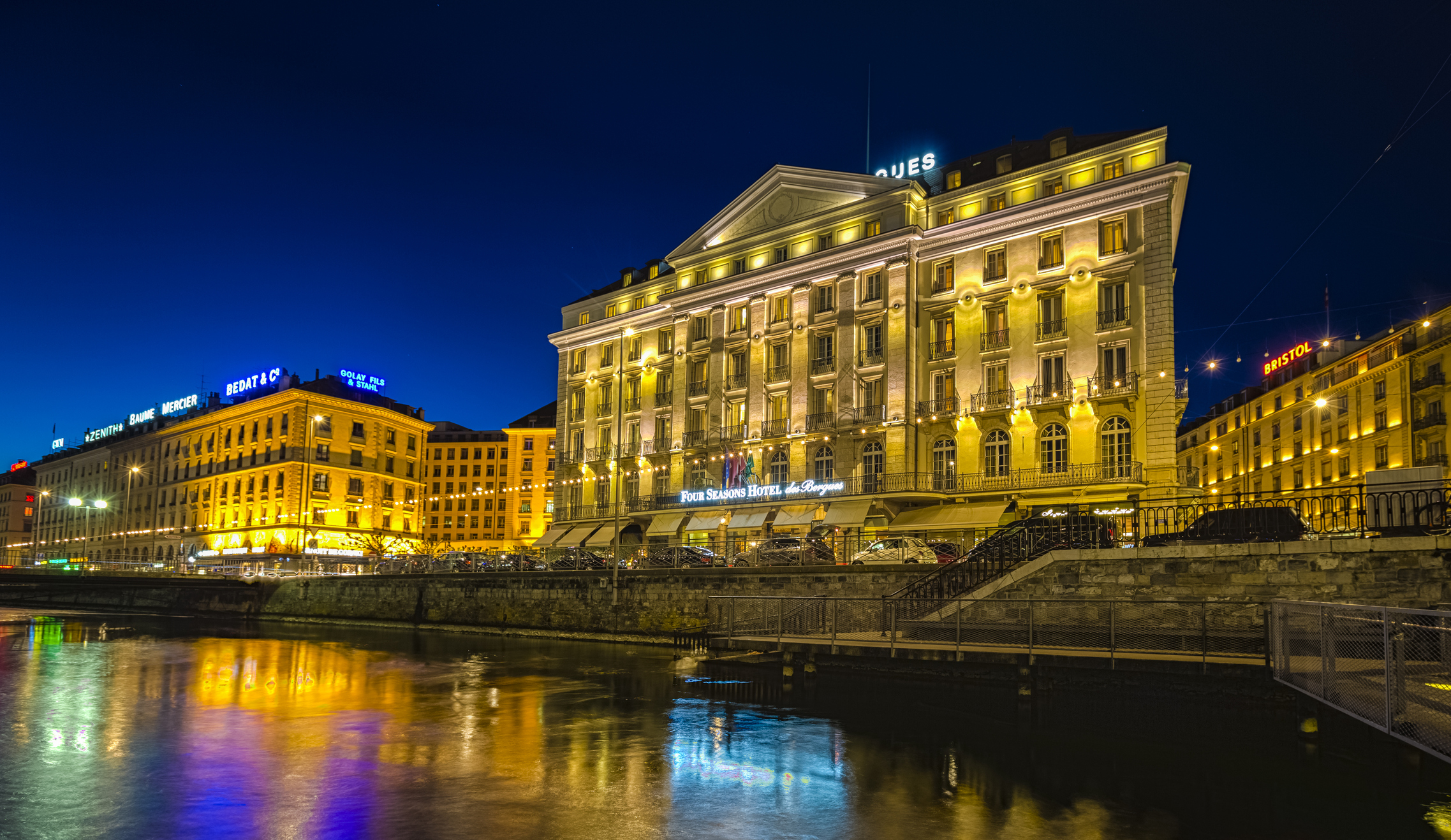 Evening view of a brightly lit hotel by a river with reflections on the water