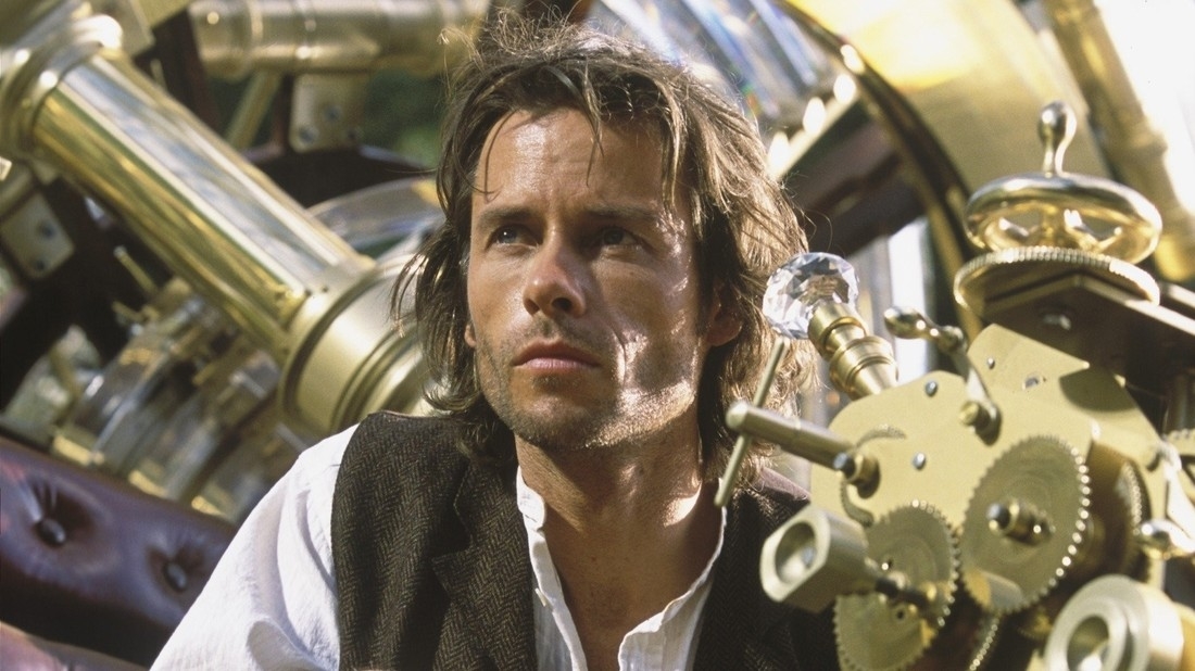 Man with disheveled hair in a vest, sitting by intricate machinery, looking pensive