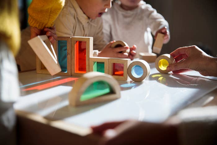 Children playing with wooden blocks and shapes on a table, with an adult&#x27;s hand visible