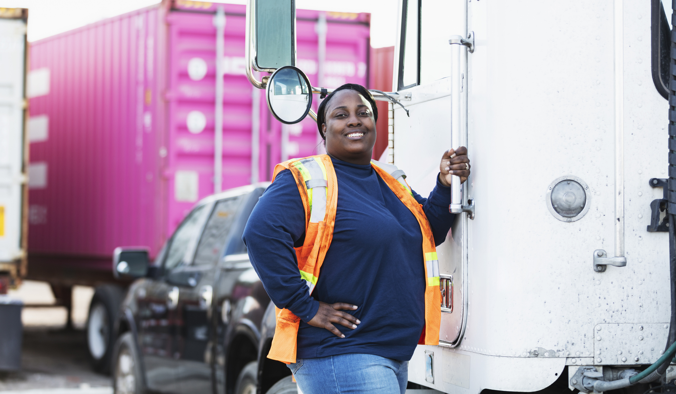 A confident worker in a safety vest stands by a truck, hand on hip, promoting women in transport