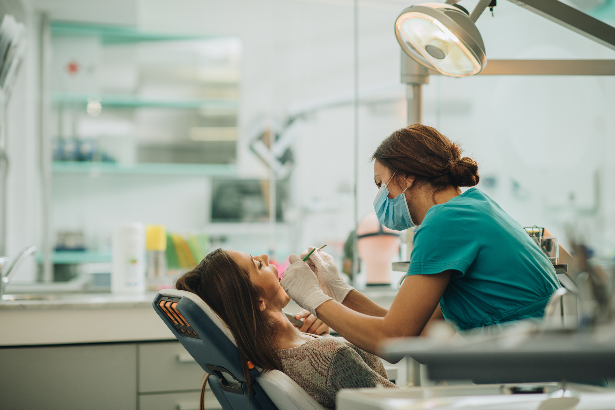 Dentist in scrubs examines a patient in a dental chair under an overhead light